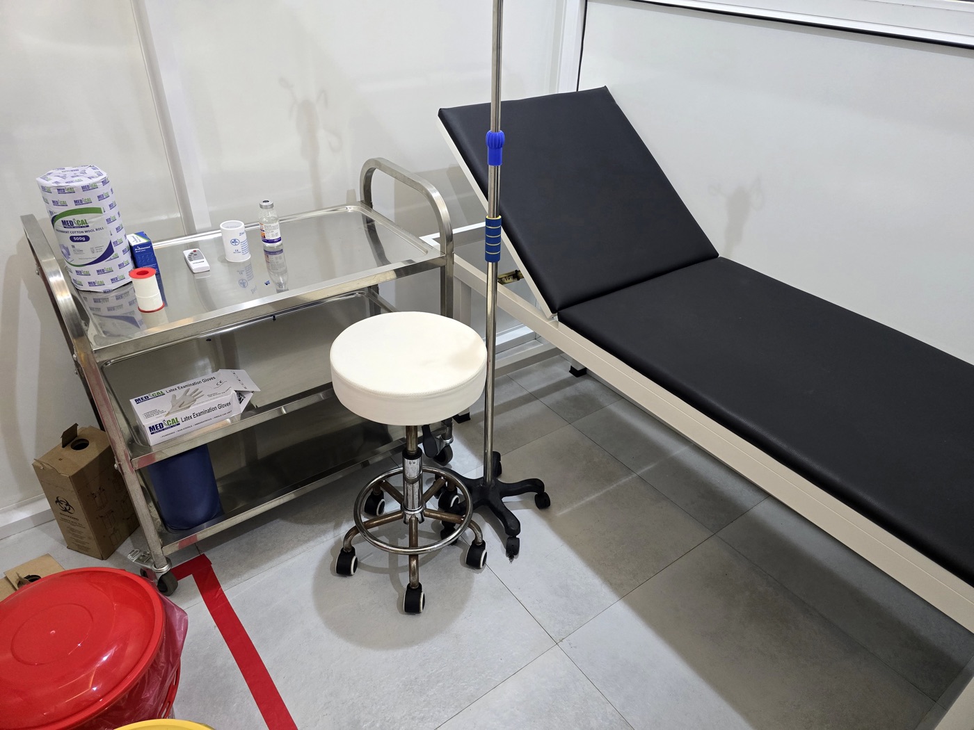 A treatment room at MacWish Medical Centre, with an examination couch, a stool, a drip stand and a dressing trolley